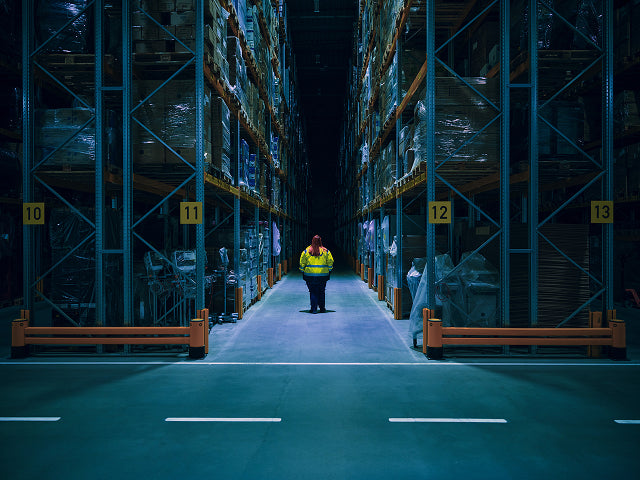 Person in a warehouse wearing a high-visibility jacket walking between rows of shelves.