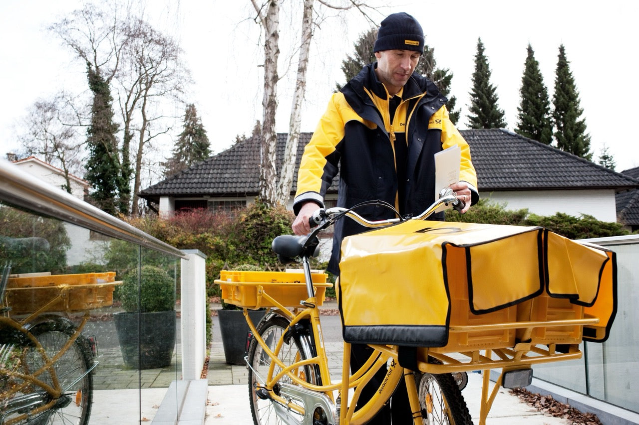 Deutsche Post delivery person on a yellow cargo bike with a large bag, in an urban setting.