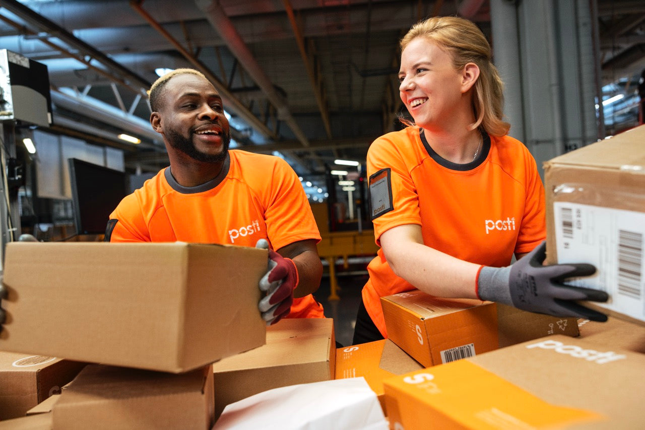 Two Posti workers in orange uniforms packaging boxes in a warehouse.
